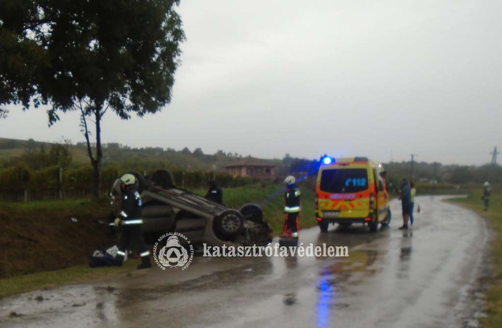 fejtetőre borult autó a vizes út szélén, tűzoltók, mentőautó