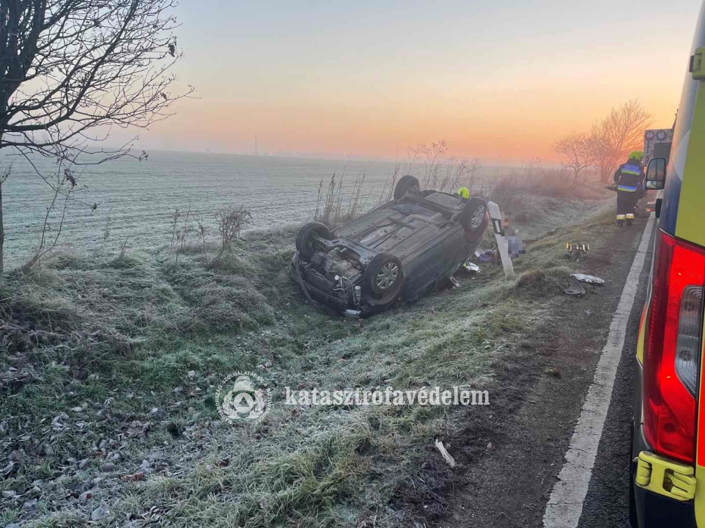 fejtetőre borult autó az árokban, mögötte tűzoltók, előtérben mentőautó